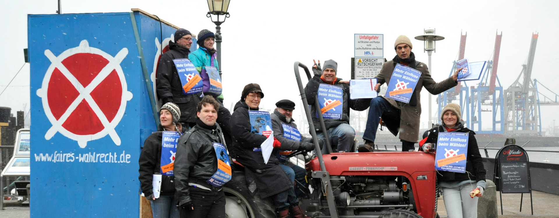 Auf dem Bild sind 9 Menschen auf einem Traktor, jeder hat ein kleines Plakat mit der Aufschrift Mehr Einfluss beim Wählen und im Hintergrund sind Hafenkräne zu sehen und zwei Warnschilder mit der Aufschrift; Unfallgefahr auf Brücken und Ponton und Vorsicht Rutschgefahr