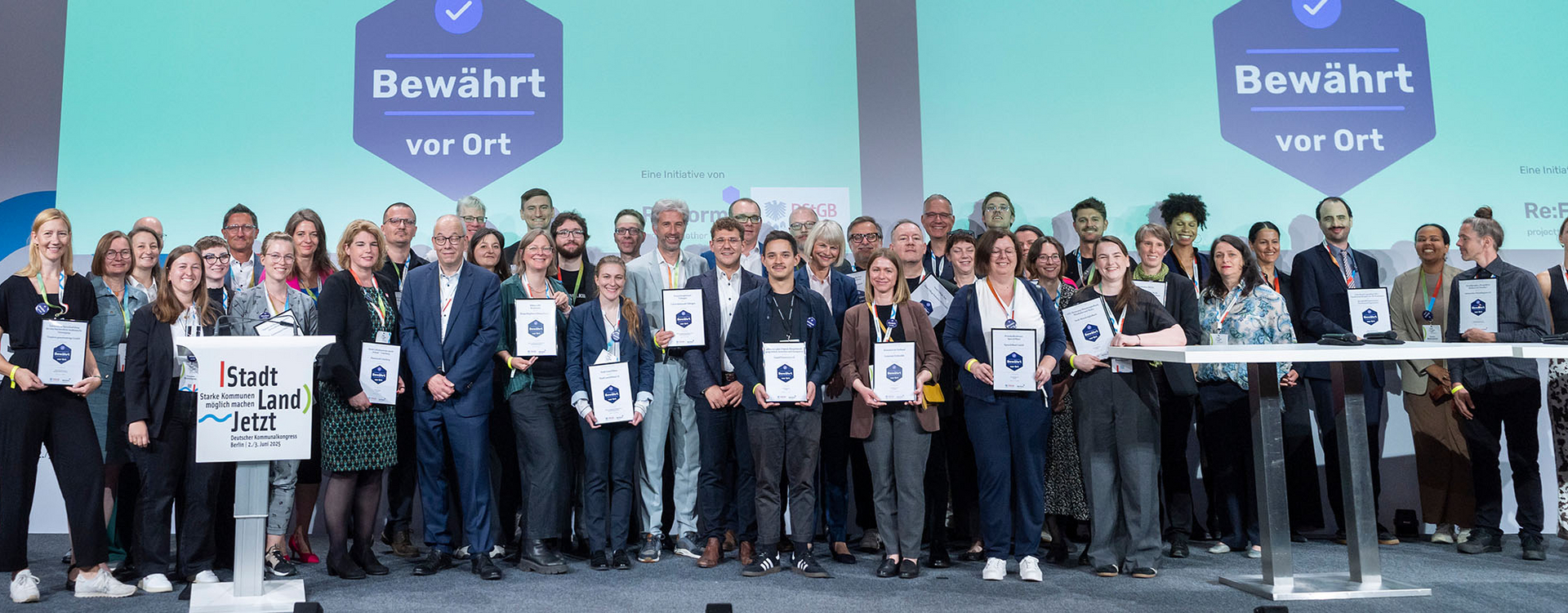Circa 45 Menschen stehen auf einer Bühne und manche halten ein Blatt hoch.