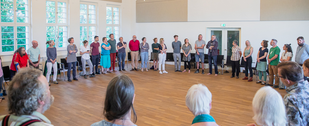 40 Menschen stehen im Kreis in einem Saal