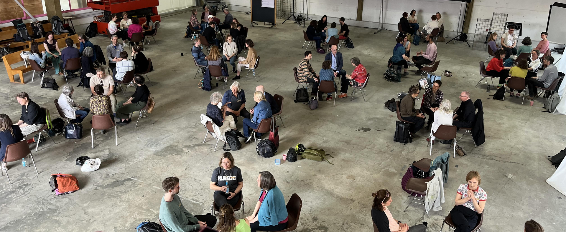 Es ist eine Industriehalle zu sehen mit Menschen, die in vierer Gruppen reden oder diskutieren. An der Wand ist von einem Beamer an die Leinwand "Das Format Sprechen & Zuhören" und weiter Text darunter abgebildet.